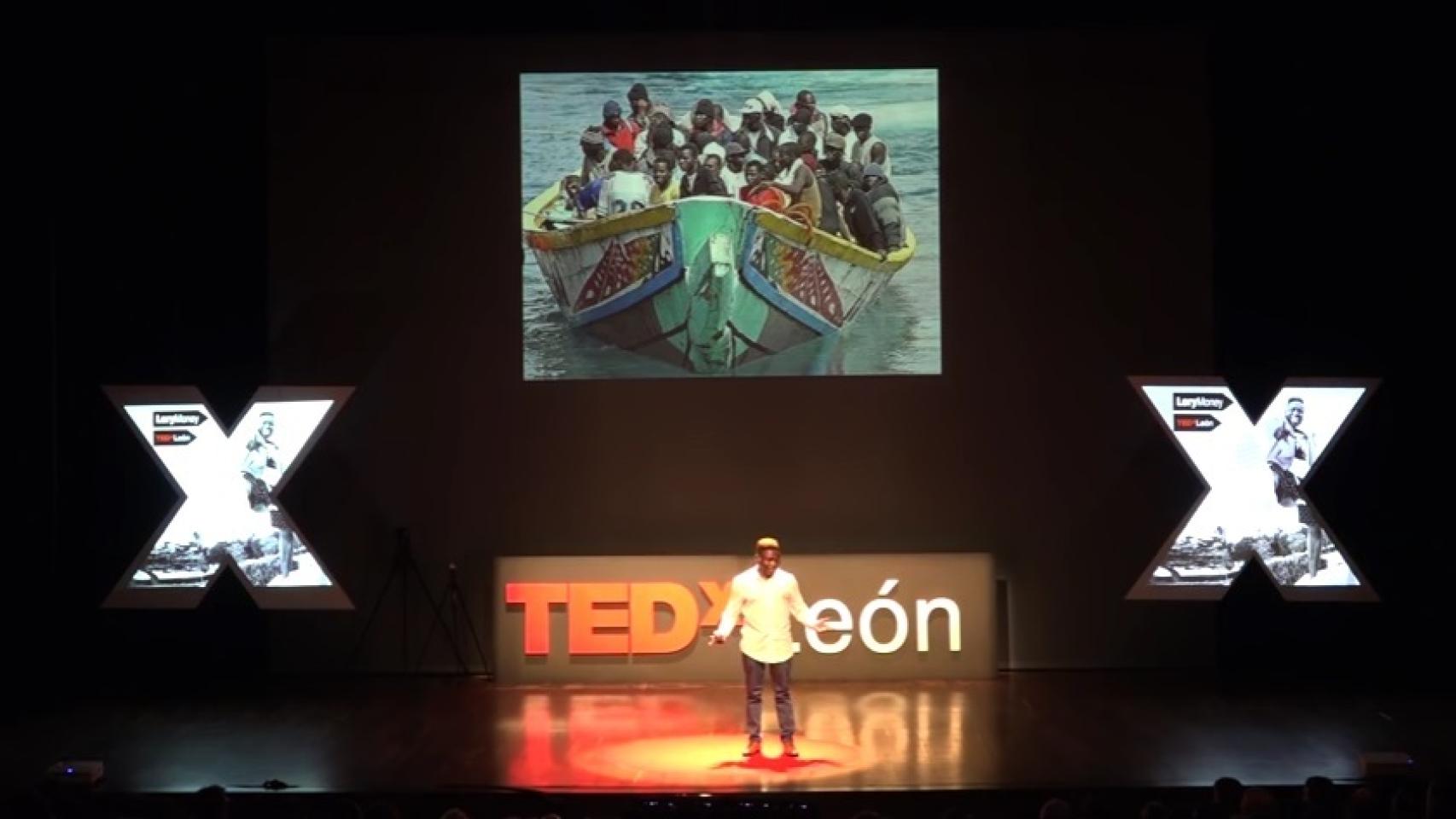 Lory Money en el centro del escenario con la foto de una patera detrás.