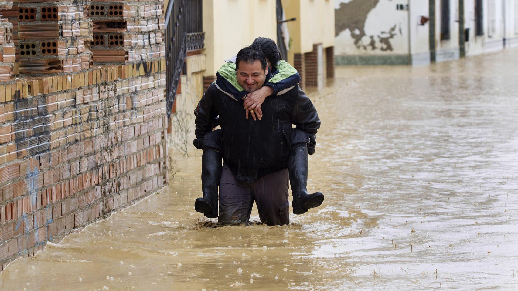 Una persona camina con otra a hombros entre las casas inundadas en la barriada Doña Ana de Cártama.