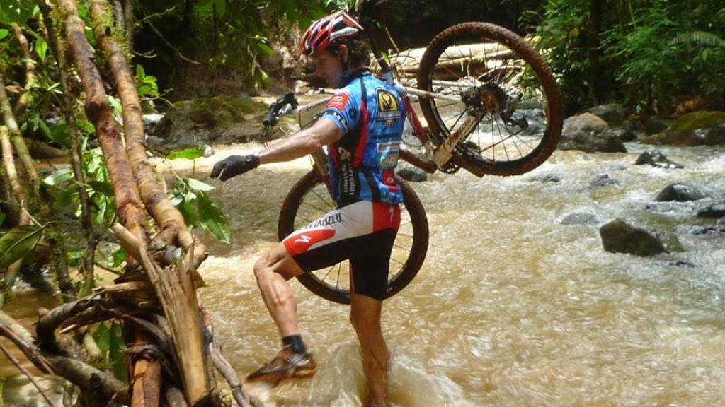 Un corredor porta a hombros su bici para cruzar un río.