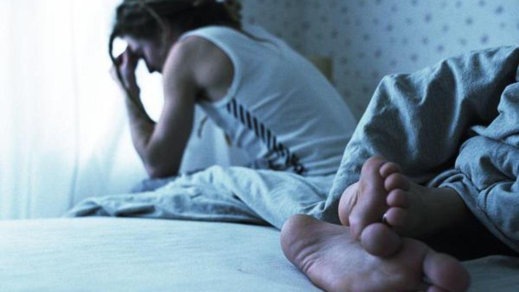 Un hombre duerme mientras su pareja reflexiona en el borde de la cama.