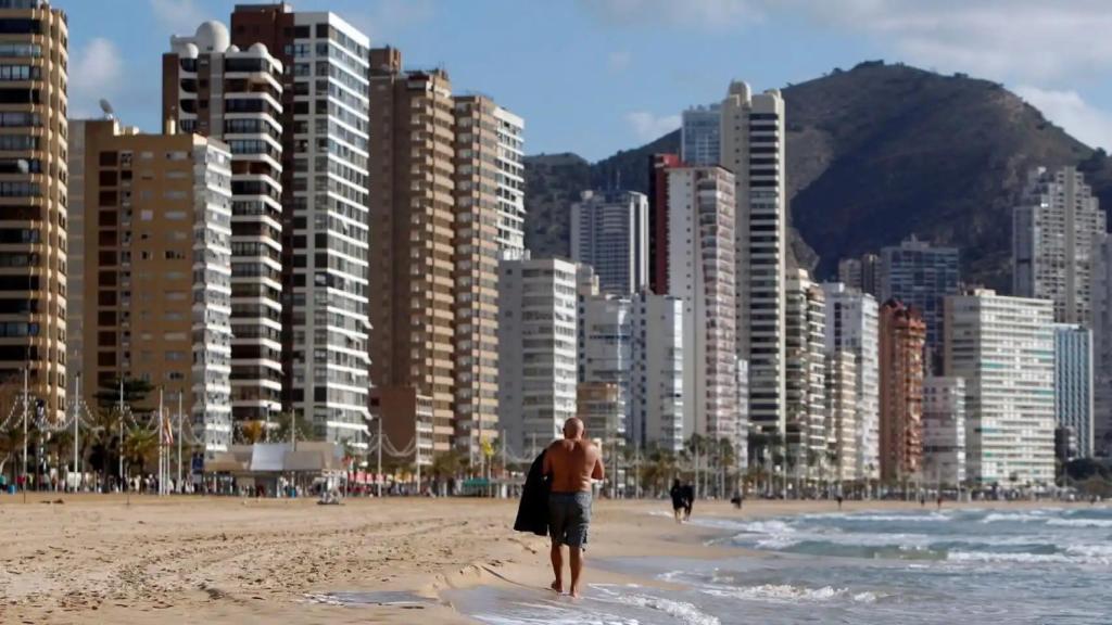 Un turista pasea por la playa de Benidorm con varios edificios de apartamentos al fondo, en imagen de archivo.