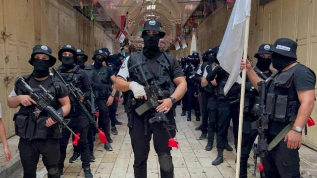 Miembros de Lions’ Den vestidos de negro, enmascarados y con las armas en alto.