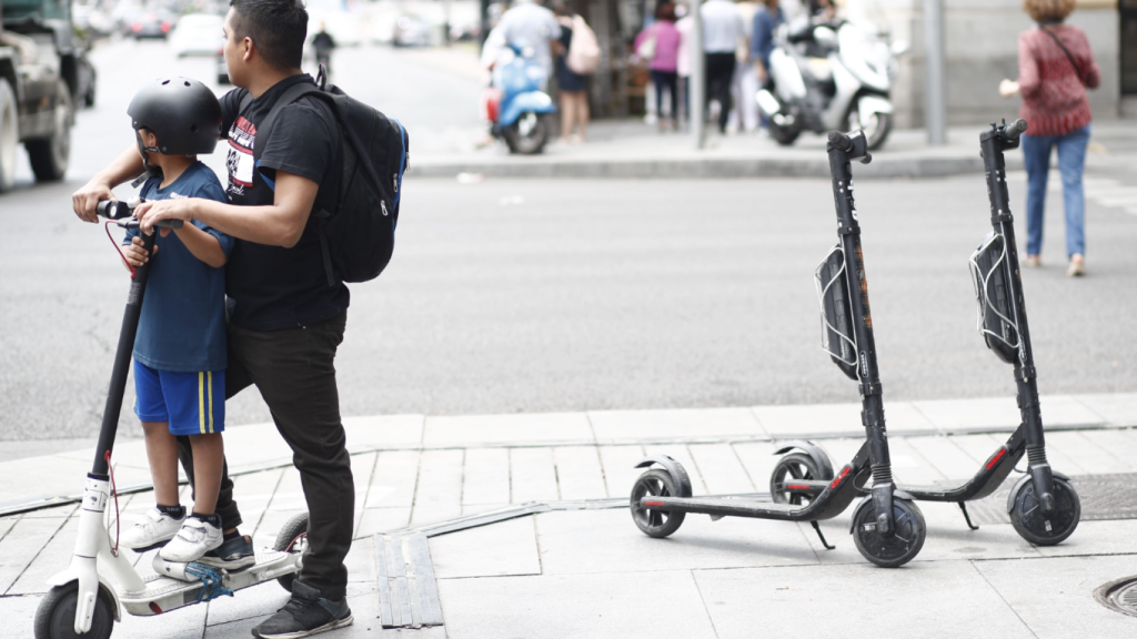 Un menor se sujeta a un patinete eléctrico con la ayuda de un adulto.