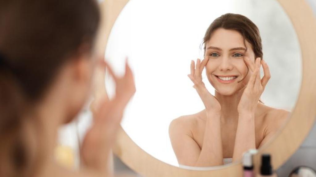 Mujer joven aplicándose crema frente al espejo.