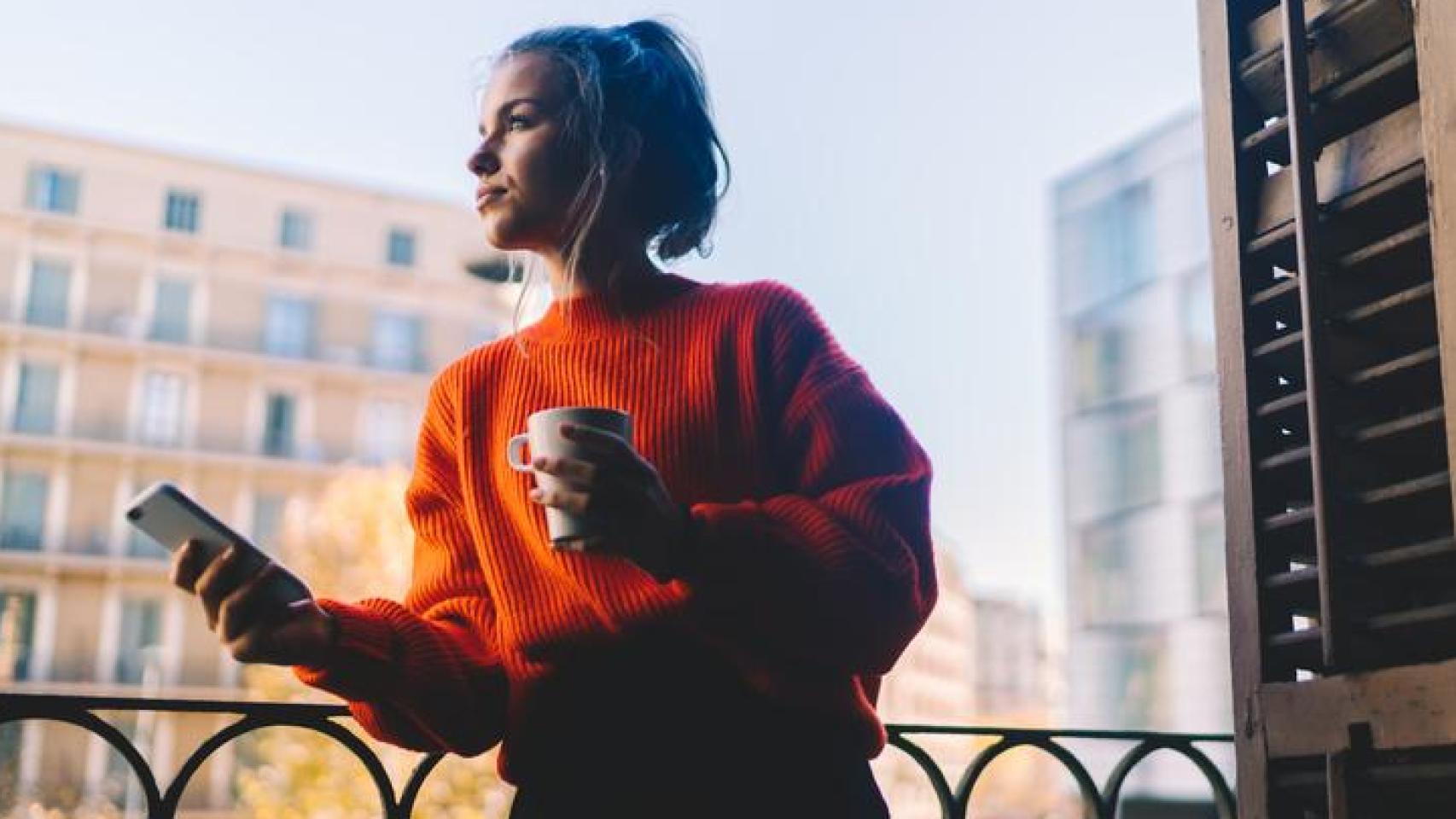 Mujer joven en el balcón con el móvil y un café.