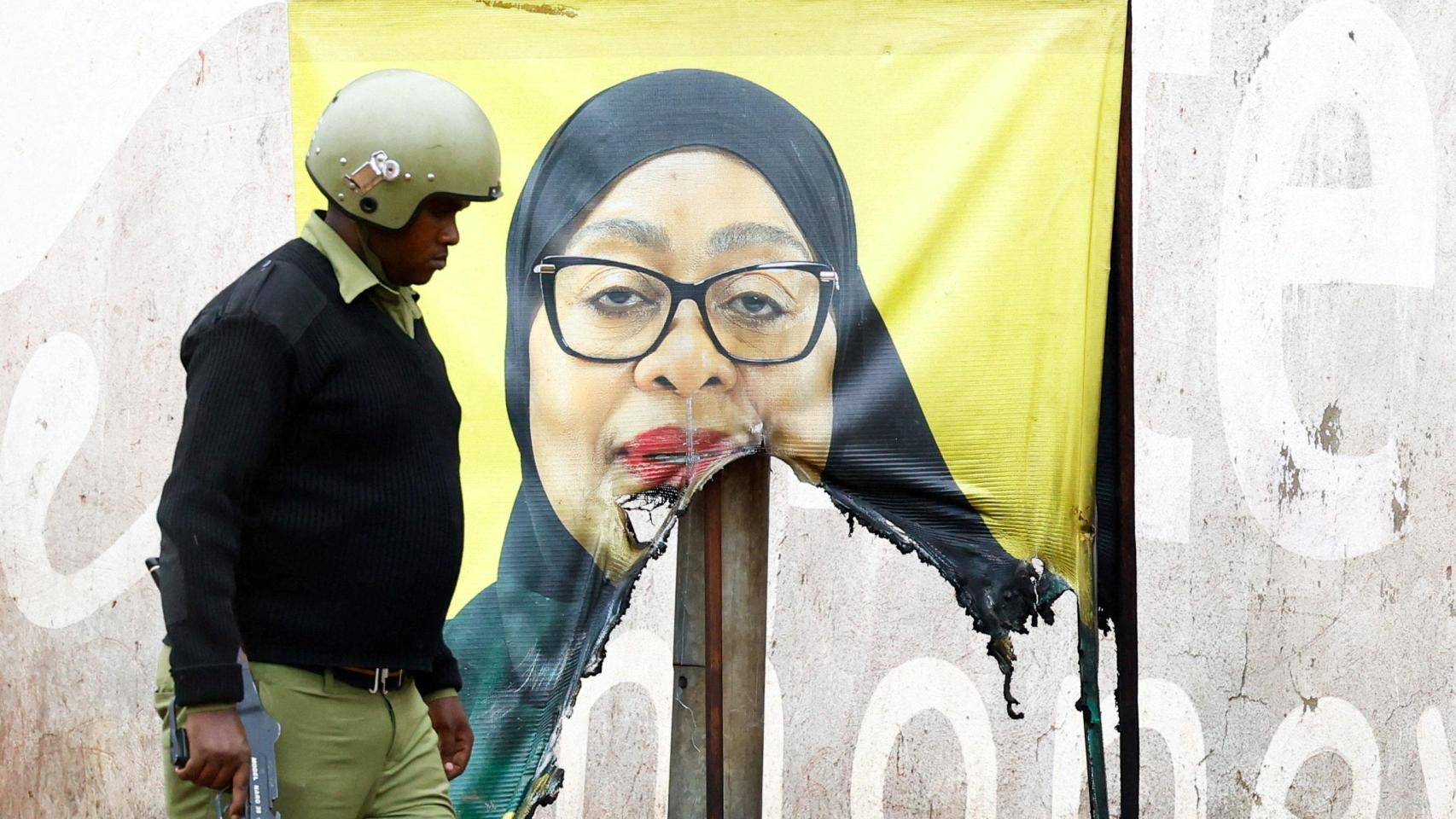 Un agente de la policía antidisturbios de Tanzania pasa junto a un cartel de campaña vandalizado de la presidenta Samia Suluhu.