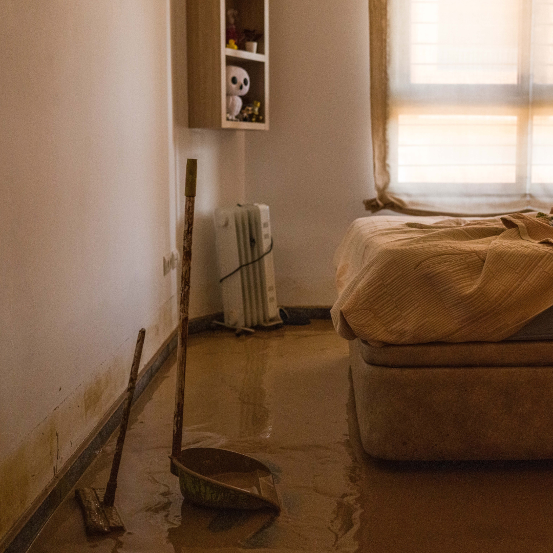 Habitación de vivienda tras una DANA: suelo cubierto de barro y agua estancada, marcas de inundación en las paredes, cama deshecha elevada sobre somier, fregona y recogedor embarrados en primer plano, y luz tenue entrando por una ventana con cortinas húmedas.