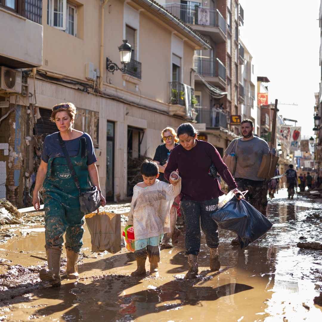 Vecinos y voluntarios caminan por una calle convertida en lodazal, con botas y ropa manchada, llevando bolsas y ayudando a un niño tras la dana en Valencia