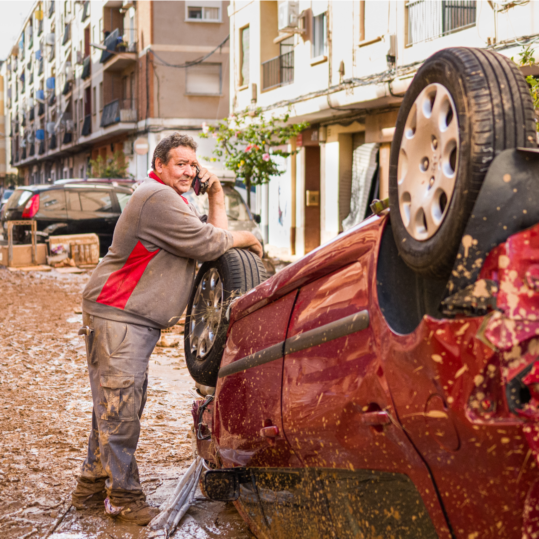 Hombre cubierto de barro habla por teléfono apoyado en un coche rojo volcado y salpicado de lodo en una calle residencial devastada por la dana en Valencia