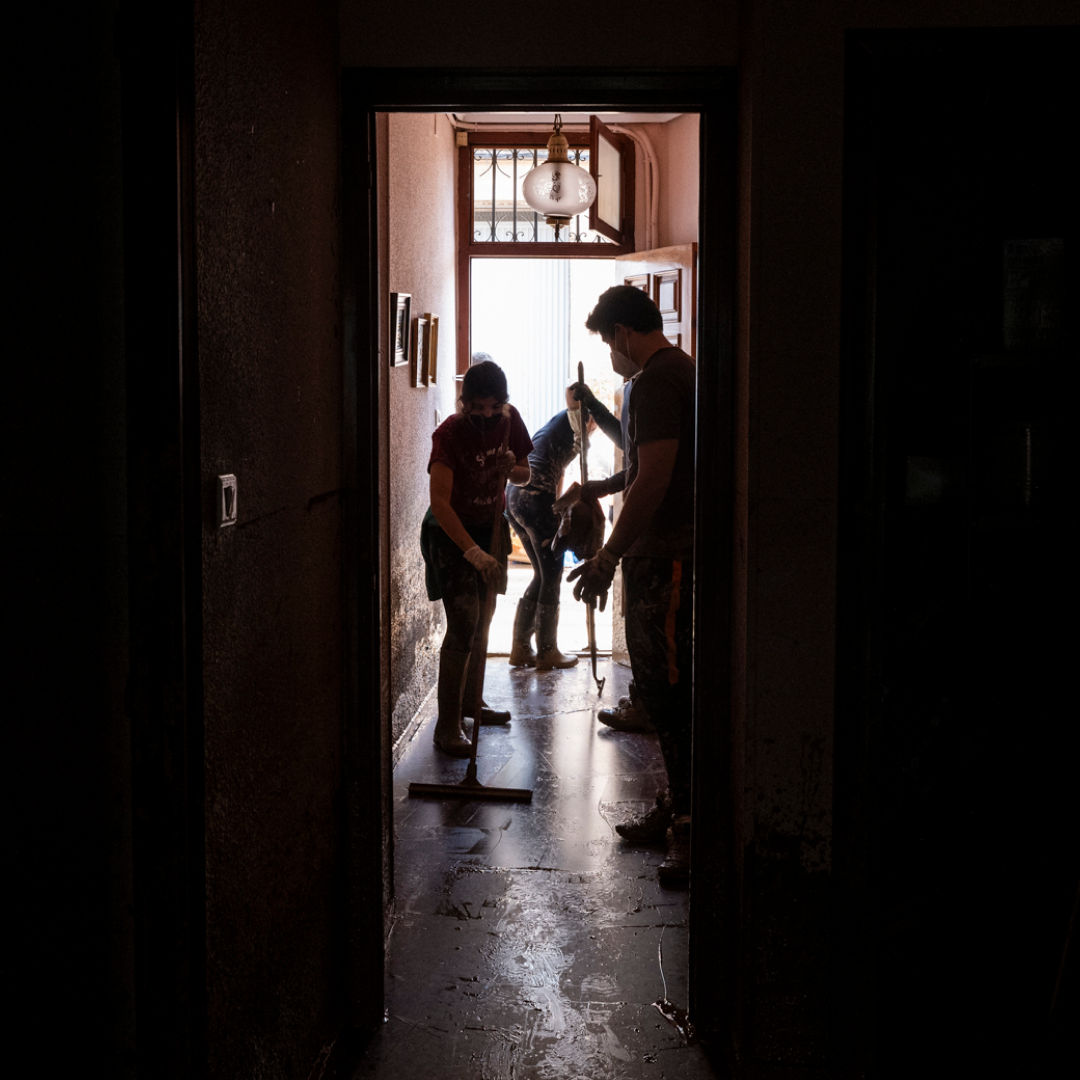 Vecinos y voluntarios caminan por una calle convertida en lodazal, con botas y ropa manchada, llevando bolsas y ayudando a un niño tras la dana en Valencia