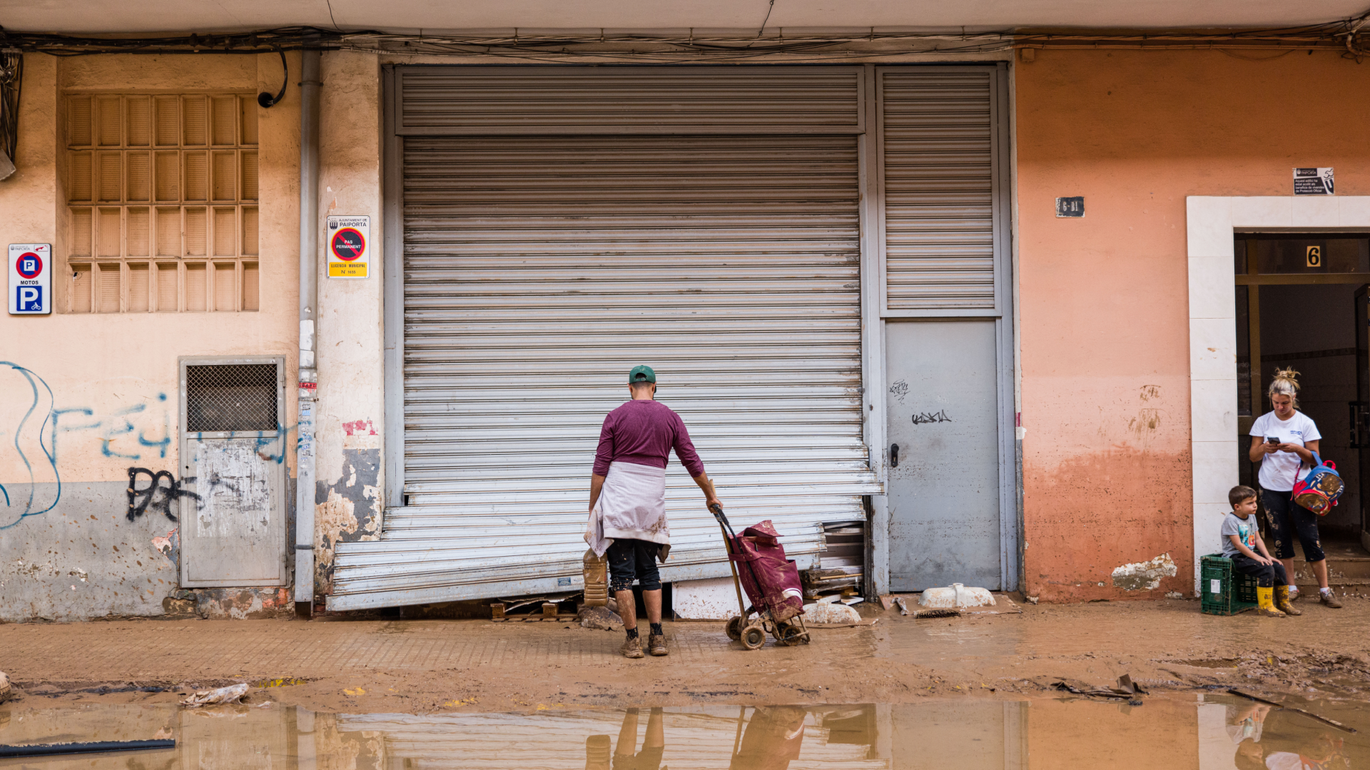 Comercio con persiana metálica abollada y barro en la acera tras la dana en Valencia; un vecino empuja un carro de limpieza frente al local mientras, a la derecha, una mujer y un niño con botas esperan en el portal entre charcos