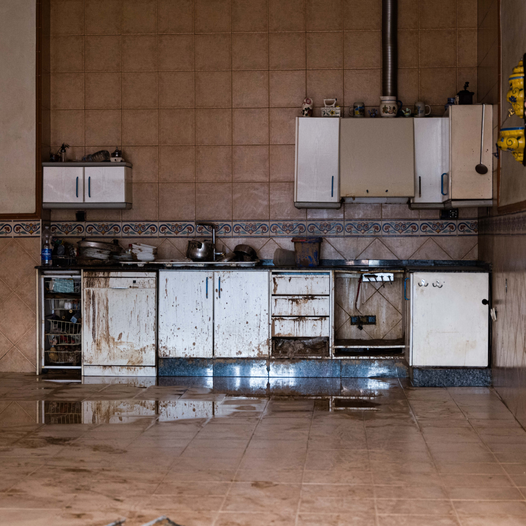 Cocina de una vivienda anegada por la DANA: muebles bajos y electrodomésticos manchados de barro, cajones rotos y charcos reflejados en el suelo de baldosas