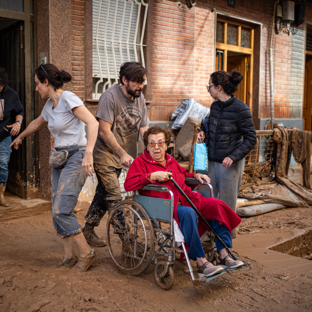 Vecinos empujan una silla de ruedas con una mujer mayor por una calle cubierta de barro frente a fachadas dañadas tras la dana en Valencia