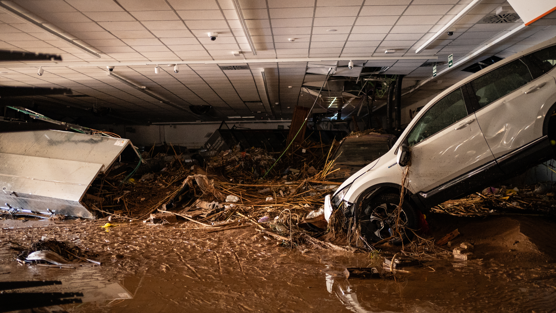Interior de un edificio inundado con techo derrumbado, montones de escombros y cañas, y un coche blanco encallado entre el barro dentro de un aparcamiento o nave tras la dana en Valencia
