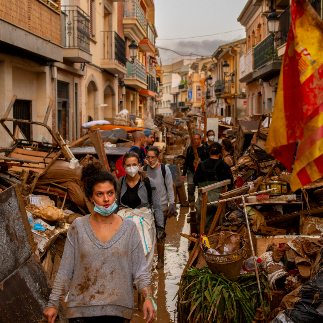 Voluntarios y vecinos caminan por una calle estrecha anegada y flanqueada por montones de muebles y escombros cubiertos de barro tras la dana en Valencia