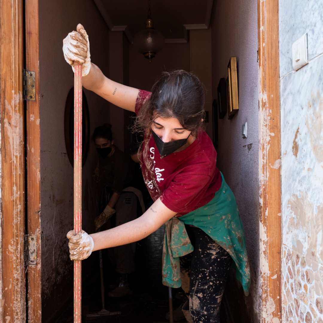 Joven con mascarilla y guantes limpia con una escoba el barro del interior de una casa, con ropa manchada y paredes salpicadas tras la dana en Valencia